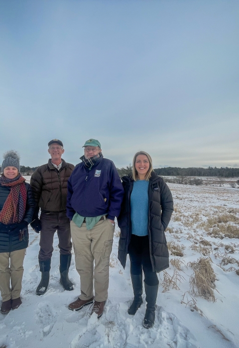 Four people stand in the foreground, with snowy land, cloudy sky, and a large green tree behind them