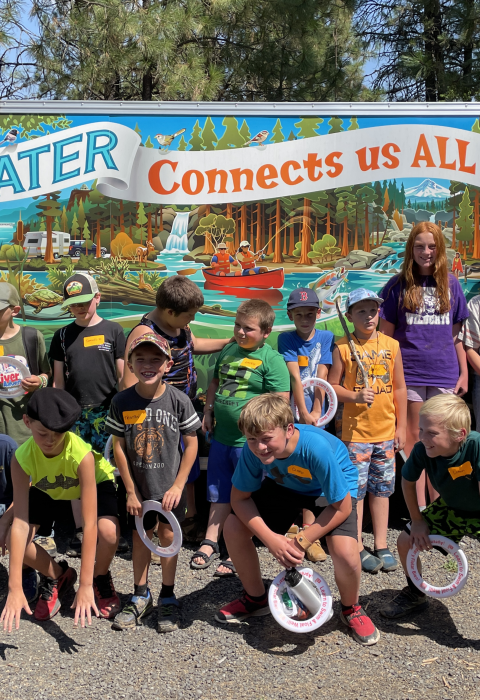 Several kids pose in front of a colorful mural with a banner that says, "Water Connects us ALL"