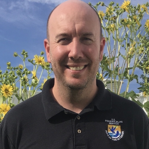 Color photo of Daniel Elbert wearing a black polo shirt with US Fish and Wildlife Service emblem on the chest. Daniel has sunflowers in the background and is smiling.