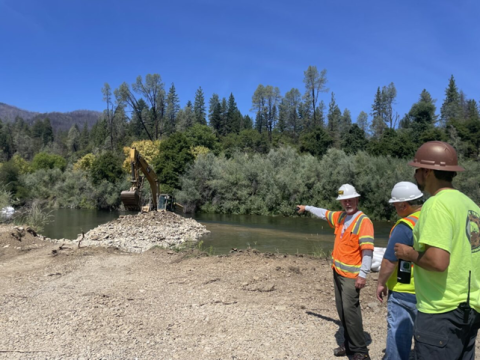 Three people view heavy machinery work being conducted along a riverbank. 