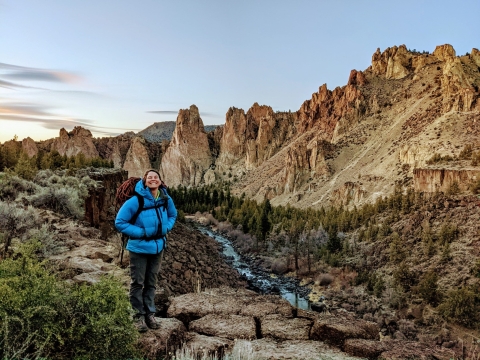 A woman wearing a blue puffy jacket and backpack stands smiling in front of a scenic view of rock cliffs and a river.