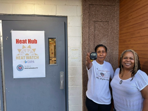 Image of 2 volunteers in front of a door labeled Heat Watch, one volunteer holding up a thermometer