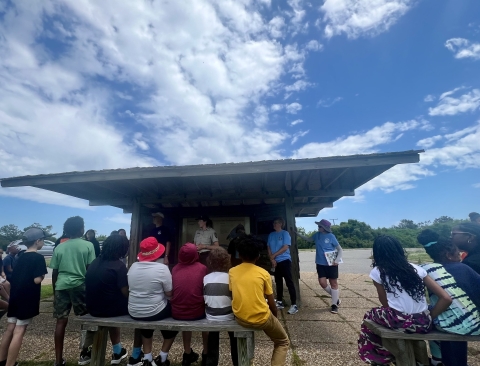 Students sit on benches in front of a line of wildlife refuge staff and volunteers. The group is gathered around a covered kiosk. The sky overhead is blue with puffy white clouds.