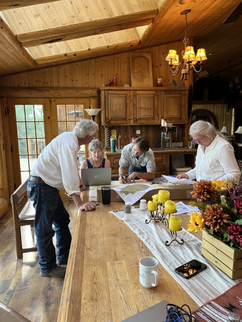 The biologist is sitting at a computer looking at the screen while three partners circle around her. One partner is looking at the computer screen and two others are hovered over a large paper map which is on the table next to the biologist.