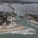 image shows an island neighborhood and houses connected to mainland by a bridge, which has been washed out by water. Debris litters the shoreline