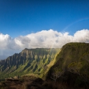 A view of mountains on Kauai where the mitigation site is. The clouds break behind the mountain cliffs and a rainbow arches in front.