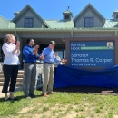 a group of people stand at a large visitor center facility. One of the people pulls off a blue fabric to unveil a sign that says "Bombay Hook Senator Thomas R. Carper Visitor Center". People are clapping in celebration of the event.