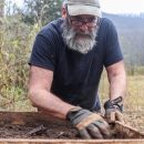 Man standing on the edge of a field, wearing work gloves, and sorting through dirt in a large sieve in front of him