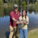 Service staff member and youth event participant posing with a rainbow trout. Lake, houses and trees in the background.