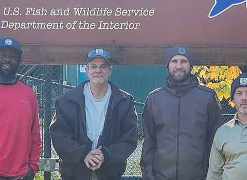 Four men stand smiling in front of a large sign for the Eagle Creek National Fish Hatchery. Sunlight and green grass are behind them.