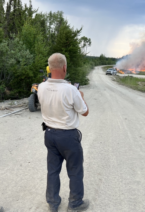 Three people watch flames burn through the forest with a truck near it along a dirt road