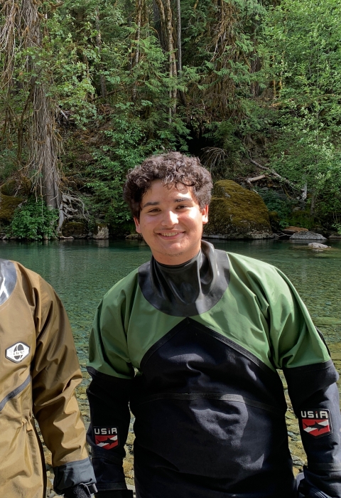 Two field crew members standing in dry suits on bank of river. River and trees in the background.