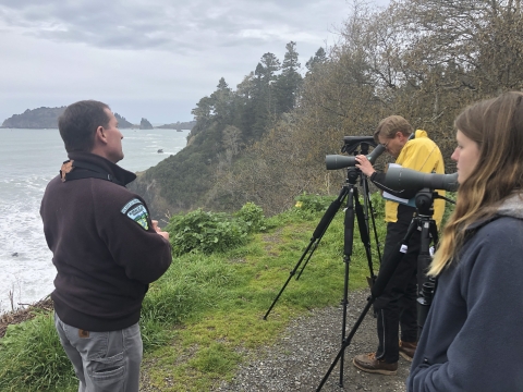 a man talking while people look through spotting scopes at the ocean.