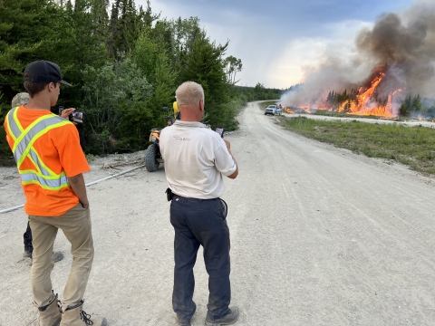 Three people watch flames burn through the forest with a truck near it along a dirt road