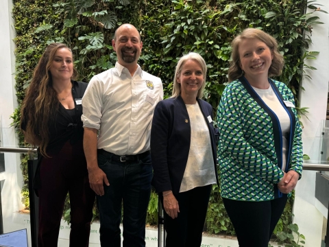 Four people stand in a line smiling for the camera. From left to right: a woman wearing a black pantsuit with long brown hair, a man wearing a white FWS-branded button up, a woman with blonde hair in a blue blazer and white shirt, and a woman with short blonde hair and a green and blue patterned cardigan over a white shirt.
