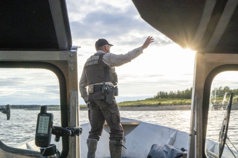 Senior Federal Wildlife Officer waves and stands at the bow of a boat while it nears the shoreline hunting camp on the Kuskokwim River, Alaska during a September moose season. 