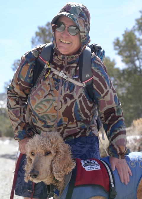 woman in hooded jacket and sunglasses smiling while petting a blonde poodle service dog, wearing a guide dog vest.