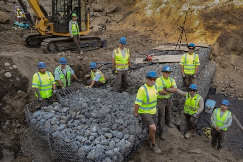 Ten people wearing bring yellow shirts and blue hard hats pose near a newly constructed rock wall