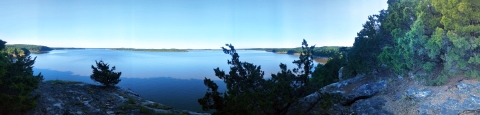 Panoramic view from lichen-covered bluff overlooking blue water and sky with green summer foliage in the distance