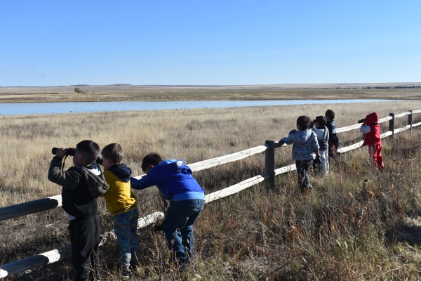 Young students standing at a wooden refuge fence looking through binoculars at a playa lake