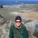 Woman in green shirt wearing stocking cap and sunglasses with brown island of and blue ocean behind her.