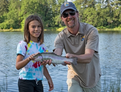 Male Service employee stands beside Big Brothers Big Sisters event participant as he holds a rainbow trout she caught. Lake in background.