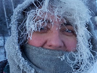 A person with frost-covered hair peeking out of a hood. Their mouth is covered by a neck gaiter and they smile with their eyes.