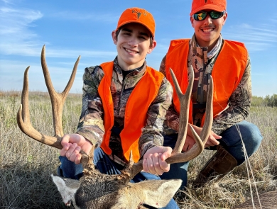 Two young brothers wearing orange and camo with the younger one holding a harvest mule deer by the antlers