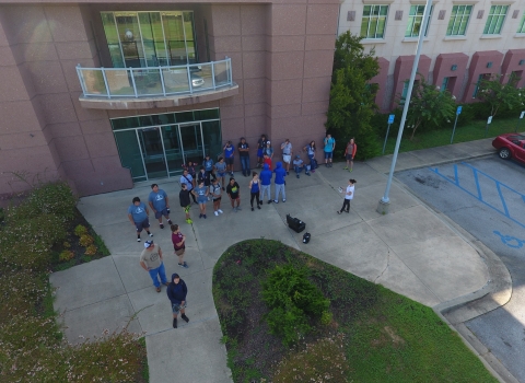 An aerial photograph of a group of people standing on the sidewalk outside of a large building