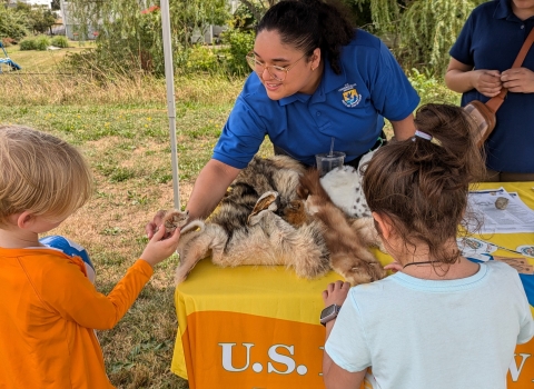 a woman in a blue USFWs uniform smiles while teaching kids about wildlife at an informational table outside