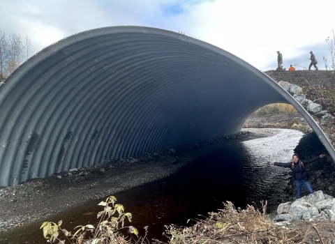 A huge culvert spanning a creek with a woman inside with outstretched arms and people walking on the road above