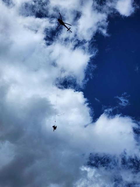 With blue sky and bright clouds as the backdrop, a firefighter dangles on a rope below helicopter.