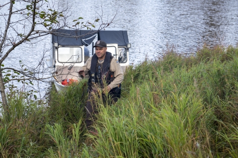 A uniformed officer walks up a river bank with a boat and river in the background 