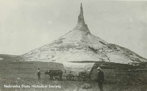 Two men, a dog, and a pair of ox with a covered wagon at Chimney Rock