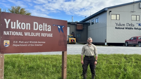 A uniformed officer stands next to a sign that reads Yukon Delta National Wildlife Refuge U.S. Fish and Wildlife Service Department of Interior. In the background is a building with large text on the side that reads Yukon Delta National Wildlife Refuge