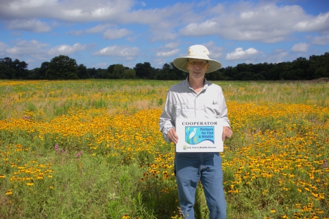 A person wearing a wide‑brimmed hat stands in a large field filled with yellow wildflowers. They hold a sign that reads “Cooperator – Partners for Fish & Wildlife – U.S. Fish & Wildlife Service.” Trees and a blue sky with scattered clouds appear in the background.
