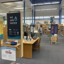 Photo of We Are Water MN Exhibit as displayed at North Branch Area Library as part of hosting experience with Chisago County. In the foreground is a welcome panel with the We Are Water Logo, as well as logos for many state partners. The background shows many other exhibit pieces at a distance, including a 12 foot tall water tower that is part of the exhibit. 