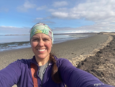 Maria Fosado smiles for a selfie on an ocean shore. Snow-covered mountains loom in the distance.