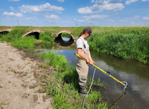 Woman in waders holds a pole and sticks it in the water. It is a summers day.