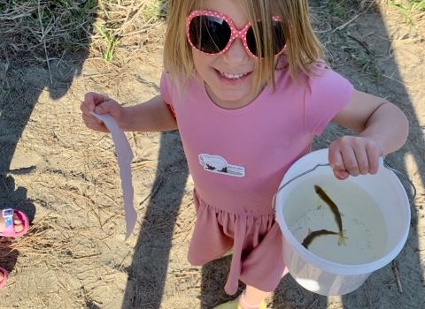 Young girl in a pink dress holds a small bucket with two fish to release