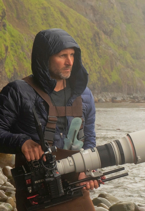 Two men carrying videography equipment walking on a rocky beach
