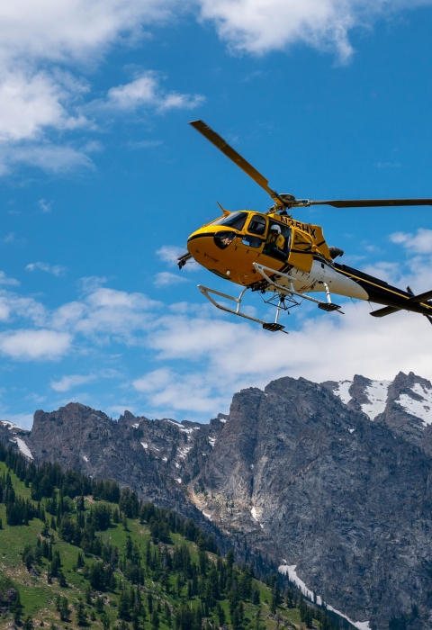 A yellow helicopter with a crewmember looking out soars above rocky mountains in Wyoming.