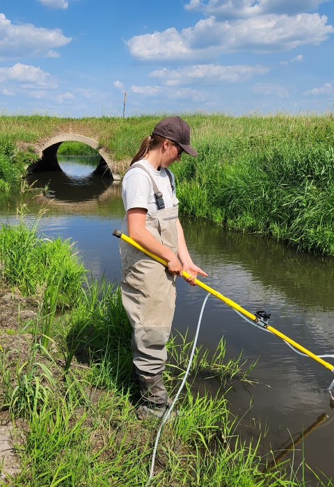 Woman in waders holds a pole and sticks it in the water. It is a summers day.