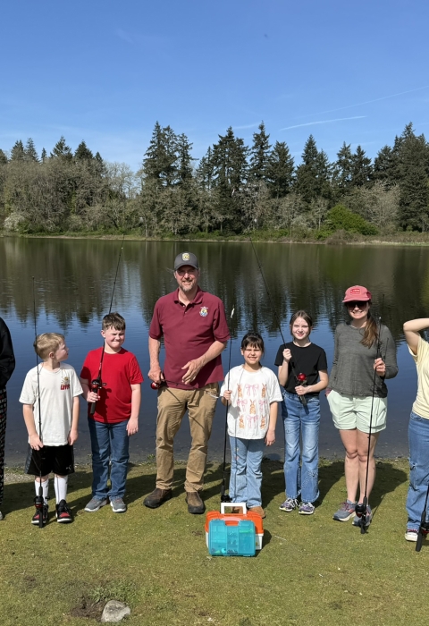 Three adults and six youth with fishing rods and tackle standing on grass in front of a lake. Lake, trees, and sky in the background.
