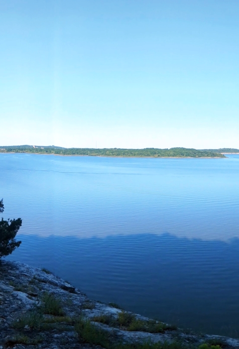 Panoramic view from lichen-covered bluff overlooking blue water and sky with green summer foliage in the distance