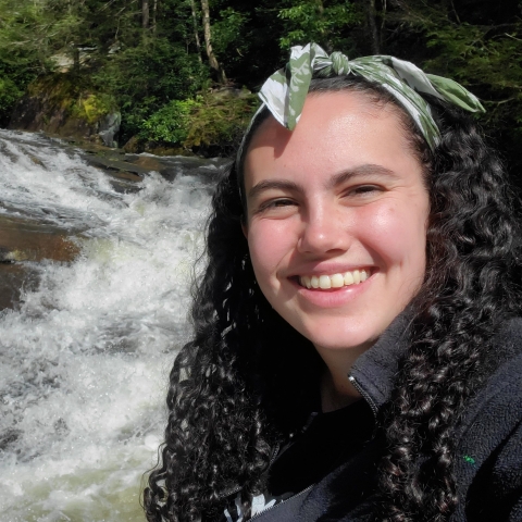 A young woman with dark curly hair smiles for a selfie in front of water rushing through a river's rapids. Dense trees and dense hemlock forest grows on the river's opposite bank.