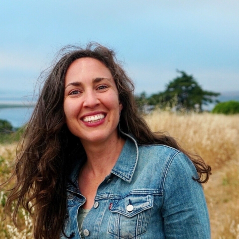 A woman smiles on a grassy hill above wetlands below. 