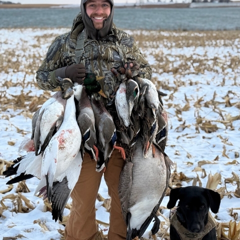 Robbie standing in a snowy field with a lake in the background and his dog at his feet holding several ducks he had hunted.