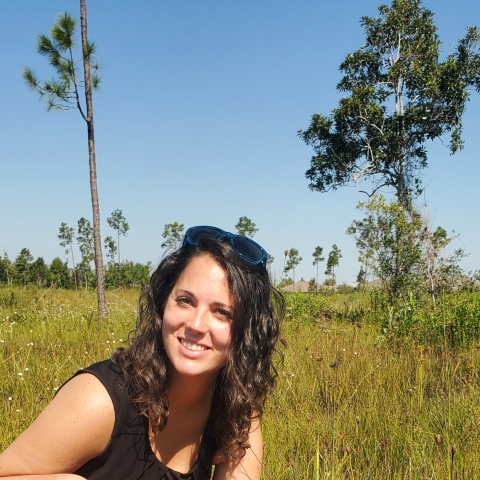 A girl kneeling by a pink flower in a field of green grass with trees in the background