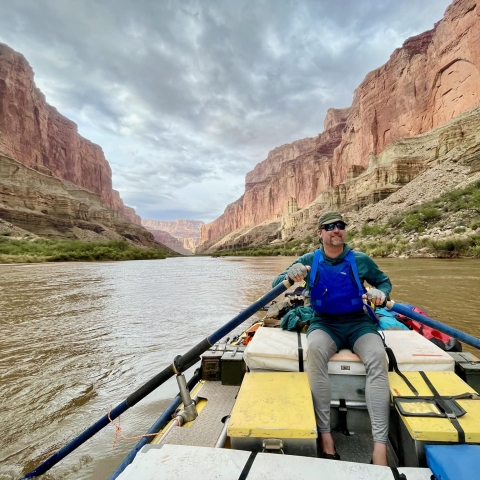 Service employee on a raft and oaring on the Colorado River. River, Grand Canyon, and cloudy sky in the background